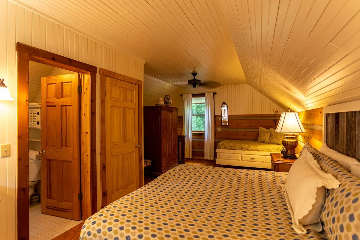 A spacious bedroom showcasing two beds—likely a king and a twin daybed—accented by wooden furniture and soft lighting from table lamps. A curtained window lets in gentle daylight, while the space feels inviting and suitable for families or couples.