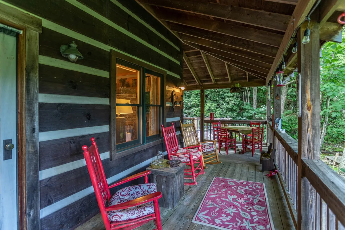 The image presents a cozy section of the wraparound porch, furnished with inviting red rocking chairs, a wooden table, and a decorative rug. The porch is surrounded by vibrant greenery, with ample space for lounging and socializing. The atmosphere is welcoming and serene, suggesting a perfect spot to sip coffee, read, or watch the sun set.