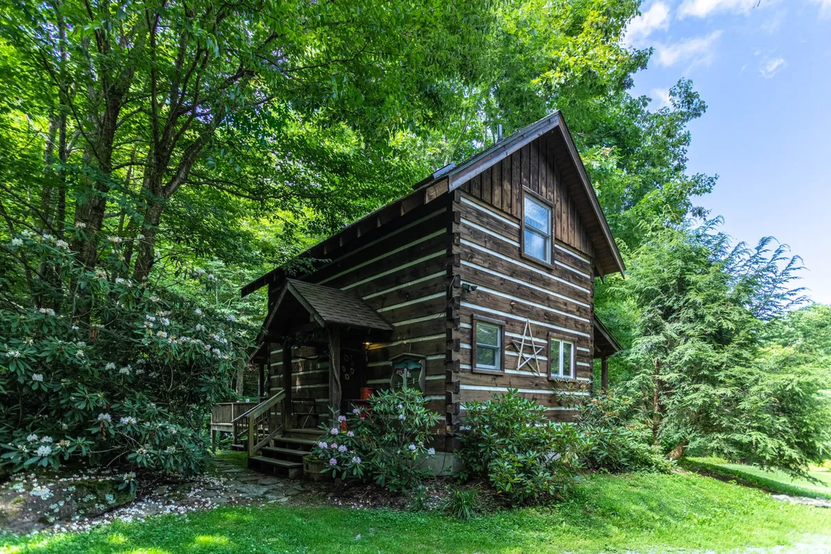 The image showcases a picturesque log cabin nestled among lush greenery, with a spacious wooden porch inviting guests to relax. The tranquil creek-side setting, surrounded by towering trees and natural beauty, immediately sets a calming, secluded mood perfect for unwinding and reconnecting with nature.