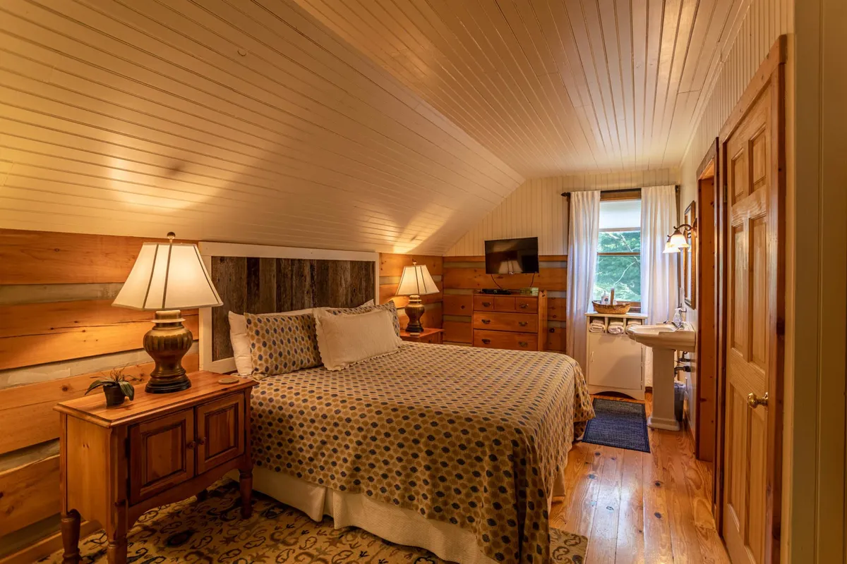 A warmly lit bedroom with a patterned king-sized bed, flanked by wooden nightstands featuring lamps. A large window brings in natural light and frames the tranquil view beyond, creating a cozy nook for morning coffee or reading.