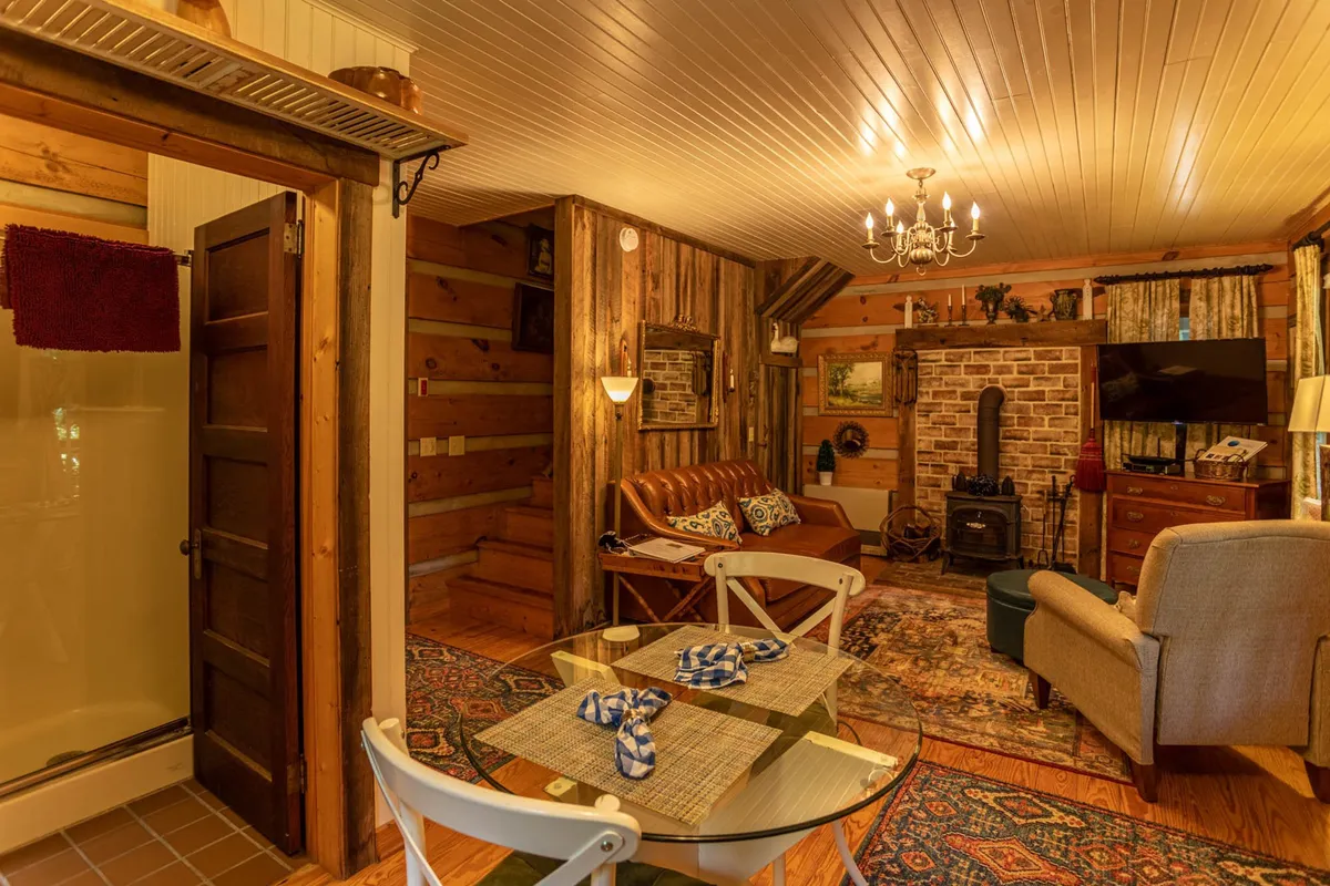 A rustic yet inviting interior scene featuring wooden walls, a brick fireplace, cozy seating, a central dining table, and a decorative chandelier. The space feels intimate and warm, with clear zones for both dining and relaxing by the fire, embodying the 'cozy comfort' described in the section.