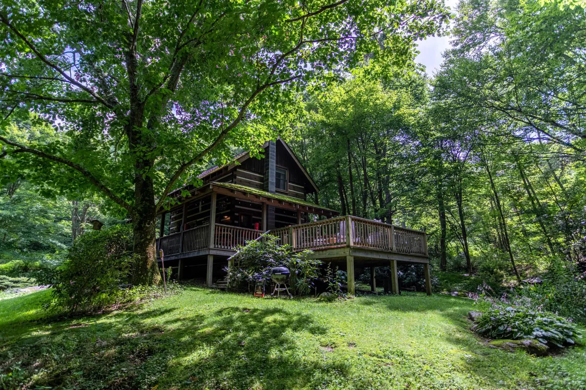 This image features the cabin nestled among tall trees, with its spacious deck extending out into the lush greenery. The serene ambiance highlights the peaceful mountain setting, with ample space for relaxing and taking in the views.