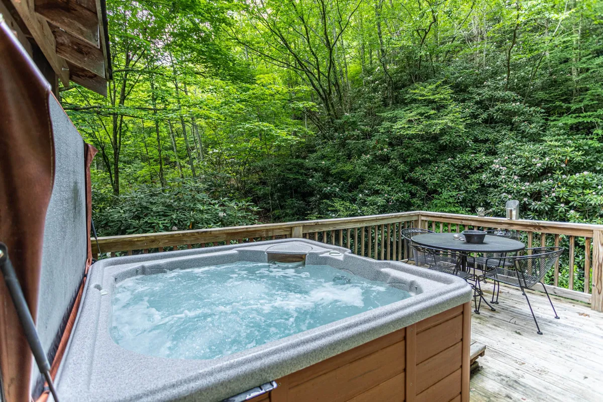 This image showcases a wooden deck featuring a hot tub with steam rising, surrounded by lush greenery. The deck is positioned above a gently flowing creek, which can be glimpsed through the trees below. Outdoor seating and a dining area invite guests to linger and enjoy the views of the mountains and water. The setting is peaceful, with clear cues for relaxation and indulgence, such as people enjoying coffee and a starlit sky in the evening.