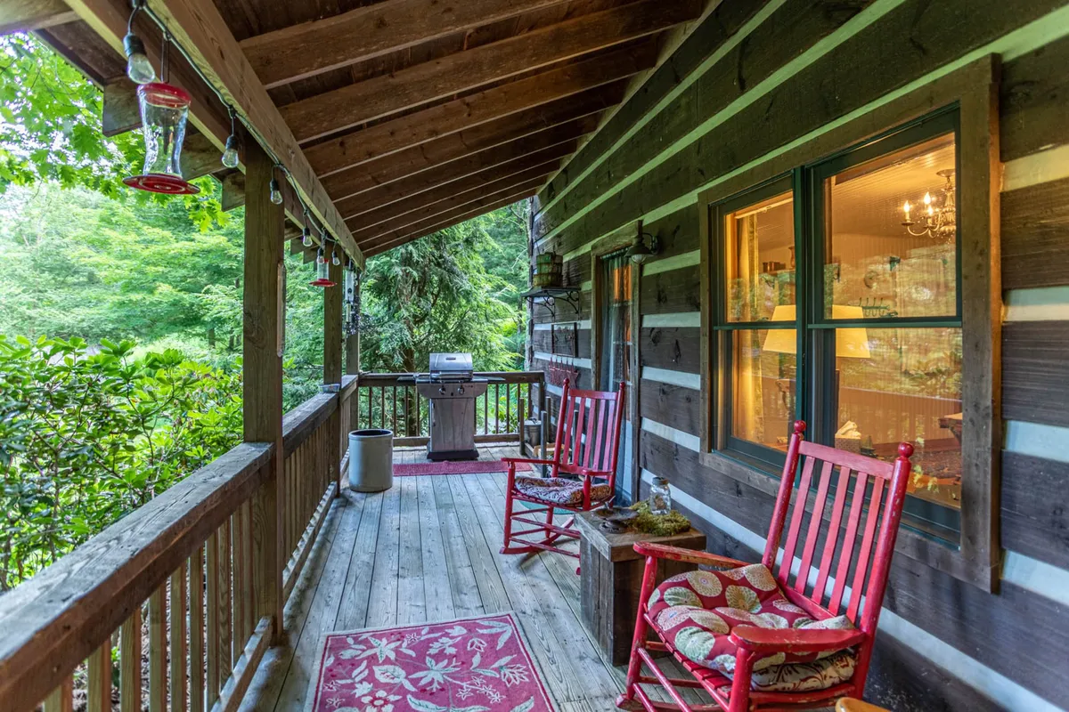 The wraparound porch is furnished with rocking chairs, a grill, a decorative rug, and wooden railing, all surrounded by verdant greenery. The view encourages relaxation, whether for morning coffee or sunset wine.